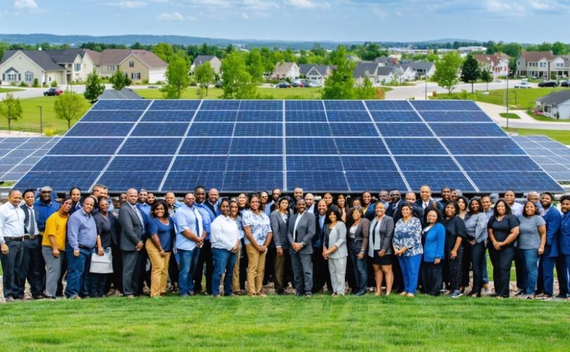 Diverse community members collaborating around a large solar panel installation, with residential neighborhoods in the background, symbolizing energy independence and community integration.