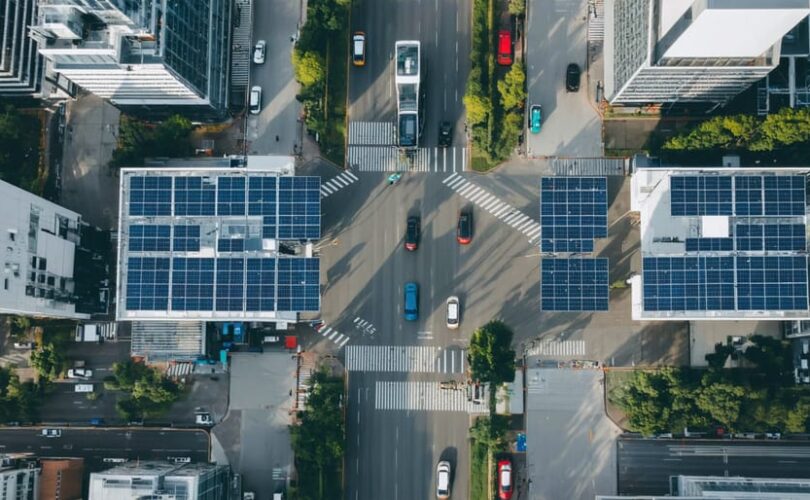 Aerial view of a cityscape featuring solar-powered electric vehicles, buses with rooftop solar panels at charging stations, and solar canopies over streets, highlighting the integration of solar energy with urban transport infrastructure.