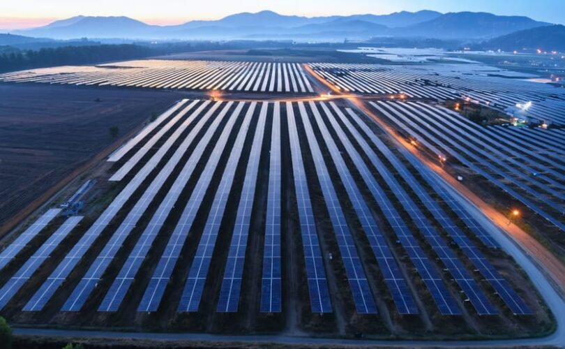 Oblique aerial view of multiple solar farms at golden hour with glowing light trails linking the arrays, suggesting optimized sequencing and routing; service roads, a substation, and low hills appear in the background.