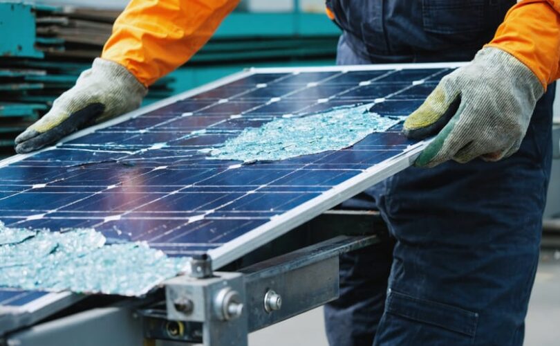 Worker in safety gear lifting a cracked solar panel onto a conveyor inside a recycling facility, with stacks of decommissioned panels and sorting machinery softly blurred in the background under diffused daylight.