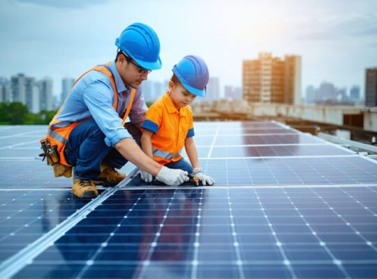 Senior solar technician mentoring a junior colleague on a commercial rooftop, both in safety gear, examining photovoltaic panels under warm golden-hour light with blurred panel rows and a distant city skyline.