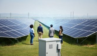 Community solar installation with rows of solar panels in a sunlit field, photographed at eye level with a few people observing the array in the distance.