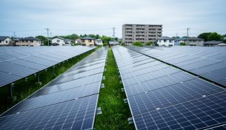 Solar panels in a community solar farm with nearby homes and an apartment building in the background under an overcast sky.