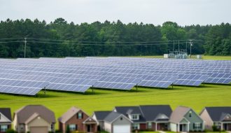 Rows of solar panels in a Georgia field with a distant neighborhood silhouette and utility lines, illustrating shared community solar access.